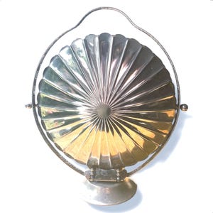 May include: A silver-toned metal serving dish with a circular, fluted shell-like design. The dish is mounted on a rotating frame with a curved handle and a circular base, allowing the central compartment to pivot on a horizontal axis.