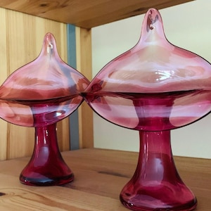 May include: Two matching, ruby-red glass decorative pieces. Each has a flared, petal-like top and a pedestal base. The glass has a glossy finish, with light reflecting off the surfaces. The pieces are displayed on a wooden shelf.