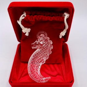 May include: A clear crystal seahorse figurine inside a red velvet box. The seahorse has a detailed, textured design and is presented against a red velvet backdrop. The box is open, revealing the figurine and a drawstring pouch.