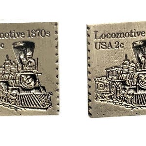 May include: Two vintage-style cufflinks with a rectangular shape and a textured, metallic finish. Each cufflink features a detailed embossed image of a steam locomotive with the text "Locomotive 1870s USA 2c" around the border.