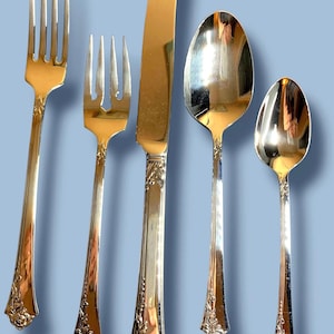 May include: A set of five silver-colored flatware pieces, including two forks, a knife, and two spoons. The utensils have a shiny finish and decorative floral detailing on the handles. The background is a solid light blue.