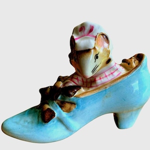 May include: A whimsical ceramic figurine featuring a mouse dressed as a chef, peeking out of a light blue high-heeled shoe. The shoe has a bow detail. The mouse wears a white chef's hat with a pink ribbon and a pink and white checkered shirt.