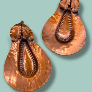 May include: Two copper pear-shaped wall hangings with textured surfaces and leaf details at the top. The interior of each pear features a textured, elongated oval design. The background is a light teal color.