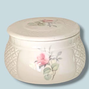 May include: A round, off-white ceramic trinket box with a removable lid. The exterior features a delicate pink rose floral motif on the front and top, complemented by a textured, embossed diamond lattice pattern on the sides of the container.