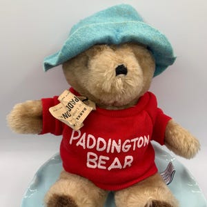 May include: A Paddington Bear plush toy wearing a red sweater with "Paddington Bear" in white lettering and a light blue hat. The bear is sitting on a light blue plate. A tag is attached to the bear.