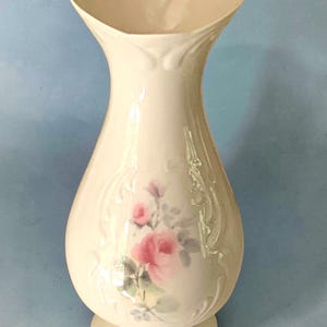 May include: A cream-colored ceramic vase with a flared, wavy rim and a rounded body. The front features a delicate floral decal of pink roses with green leaves, surrounded by embossed scrollwork patterns on the glossy, smooth surface.