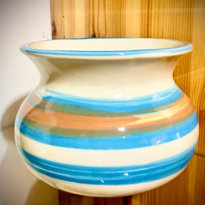 May include: A ceramic vase with a cream-colored base and horizontal stripes in shades of blue, orange, and brown. The rim and a thin band at the top are outlined in blue. The vase has a rounded body and a flared rim.