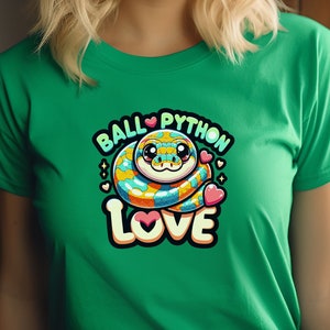 Op de afbeelding: Een groen T-shirt met een cartoon ball python die een hart draagt en de tekst "Ball Python Love" in regenboogkleuren.