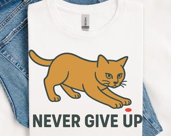 Pode incluir: Camiseta branca com um desenho de gato de desenho animado em tons de laranja e preto, alcançando um pequeno ponto vermelho. O texto "NEVER GIVE UP" está impresso em verde escuro abaixo do gato. A camiseta é exibida em um fundo jeans.