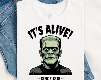 May include: White t-shirt with a green and black illustration of Frankenstein's monster, the text "IT'S ALIVE!" above and "SINCE 1818" below. The shirt is displayed on a pair of blue jeans.