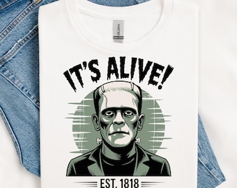 May include: White t-shirt with a graphic of Frankenstein's monster, the text "IT'S ALIVE!" above the image and "EST. 1818" below. The design is in black, white, and green. The shirt is folded on blue denim jeans.