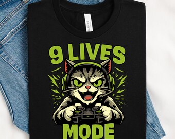 Puede incluir: Camiseta negra con un gato de dibujos animados que lleva auriculares y sostiene un mando de videojuegos. El texto "9 LIVES MODE" es verde. El gato tiene ojos verdes y un patrón de rayas grises y negras.