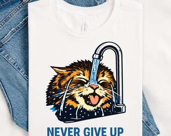 May include: White t-shirt featuring a cartoon cat with water pouring on its face from a tap. The cat is brown, orange, and black. The text "NEVER GIVE UP" is printed in blue. The shirt is displayed on a denim background.
