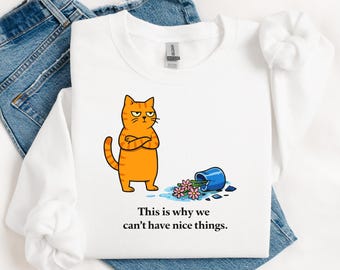 May include: White long-sleeve shirt with a cartoon orange cat with crossed arms, a spilled blue vase, and pink flowers. The text reads: "This is why we can't have nice things."