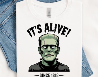 May include: White t-shirt featuring a graphic of Frankenstein's monster. The text "IT'S ALIVE!" is above the monster's head, and "SINCE 1818" is below. The shirt is displayed on a pair of blue denim jeans.