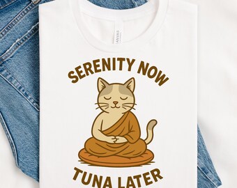 May include: White t-shirt with a cartoon cat meditating in a lotus position. The cat is tan and cream-colored, wearing a brown robe. The text "SERENITY NOW" and "TUNA LATER" are in brown, arched above and below the cat illustration.