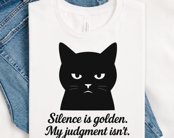 May include: White t-shirt featuring a black cat graphic with an annoyed expression. The text "Silence is golden. My judgment isn't." is printed below the cat. The shirt is displayed on a pair of blue jeans.