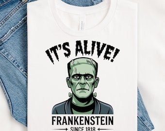 May include: White t-shirt with a green Frankenstein monster graphic, the text "IT'S ALIVE!" above the image and "FRANKENSTEIN SINCE 1818" below. The shirt is displayed on a pair of blue denim jeans.