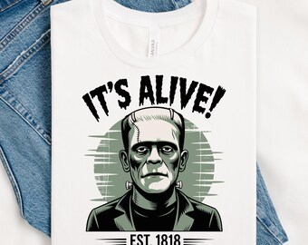 May include: White t-shirt with a graphic of Frankenstein's monster, the text "IT'S ALIVE!" above the image and "EST. 1818" below. The design is in black and green, set against a faded green circle. The shirt is folded on a pair of blue jeans.