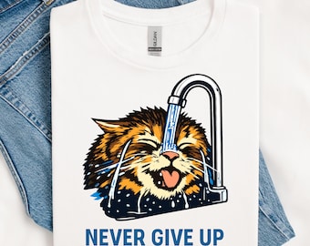 May include: White t-shirt featuring a cartoon cat with orange and brown fur, under a tap with water pouring on its head. The text "NEVER GIVE UP" is printed in blue below the graphic. The t-shirt is on a denim background.