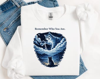 May include: White long-sleeve shirt with a graphic of a cat in a cloudscape, holding a cup. The text "Remember Who You Are" is above the graphic. The shirt is paired with blue jeans.