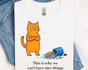 May include: A white t-shirt featuring a graphic of an orange tabby cat with crossed arms standing next to a tipped-over blue vase with pink flowers and spilled water. The text below reads, "This is why we can't have nice things."
