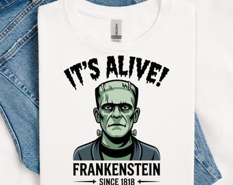 May include: White t-shirt with a green Frankenstein's monster graphic, the text "IT'S ALIVE!" above the image, and "FRANKENSTEIN SINCE 1818" below. The shirt is displayed on a blue denim surface.