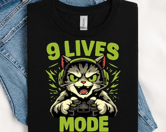 Puede incluir: Camiseta negra con un gato de dibujos animados con auriculares y un mando de videojuegos. El texto "9 LIVES MODE" es de color verde. El gato es gris y blanco con ojos y auriculares verdes. La camiseta se muestra sobre unos vaqueros azules.