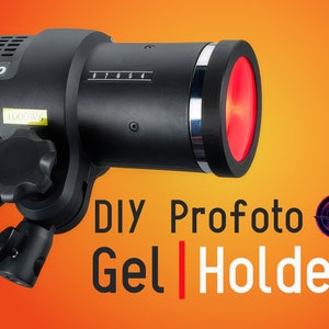 May include: A black Profoto D2 flash with a red gel filter attached to the front. The flash is mounted on a stand and has a power cord attached. The text "DIY Profoto Gel Holder" is written in white letters on a red background.