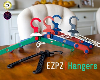 May include: Four EZPZ Hangers in different colors: green, blue, red, and white. The hangers are made of plastic and have a honeycomb design. The hangers are folded and stacked on a wooden table.