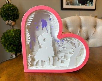 May include: A pink heart-shaped shadow box with a white paper cut-out design of a couple kissing in front of a castle. The scene is illuminated with a purple light.