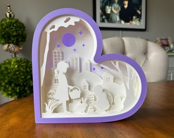 May include: A layered paper art piece in a heart shape, featuring a purple frame. The artwork depicts a girl with a basket, Easter eggs, a bunny, and a cityscape under a purple moon and stars. The overall color scheme is white and purple.