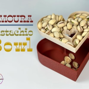 Może przedstawiać: Biała miska w kształcie serca wypełniona pistacjami, stojąca na czerwonej misce w kształcie serca. Tekst "AMOURA Pistachio Bowl" jest widoczny w tle.