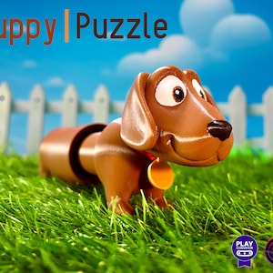 Peut inclure: Un jouet de puzzle en forme de chiot imprimé en 3D, de couleur marron et blanche, avec une expression espiègle. Le jouet est posé sur de l'herbe verte devant une clôture en bois blanc.