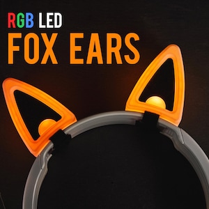 May include: RGB LED fox ears headband. The headband features two orange fox ear shapes with a glowing light inside. The text "RGB LED FOX EARS" is displayed above the ears. The headband is gray and black.