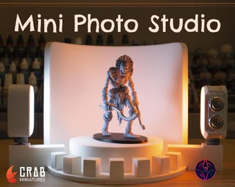 May include: A white mini photo studio with a white backdrop, two white lights, and a gray miniature figure standing on a white platform. The studio is set up on a wooden surface. The text "Mini Photo Studio" is written in white on a brown background above the studio.