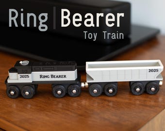 May include: A black and white toy train with a gray boxcar. The engine has the words "Ring Bearer" and "2025" printed on it. The boxcar also has "2025" printed on it. The train is a toy train.