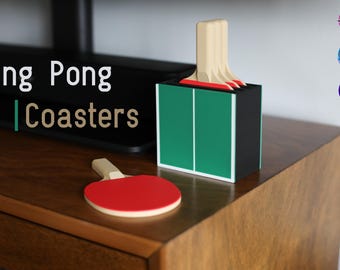 May include: A set of ping pong coasters featuring a black storage box designed like a table tennis table with green surfaces and white lines. The set includes several round red and white ping pong paddle coasters with light wood handles.