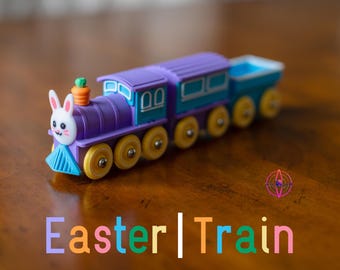 May include: A colorful toy train with an Easter theme. The train is primarily purple and teal, with yellow wheels. The front of the train features a bunny face with a carrot on top. The text "Easter | Train" is displayed below the train.