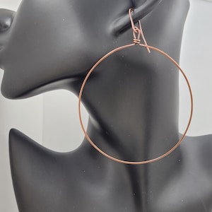 May include: A pair of large, copper-colored hoop earrings. The earrings are displayed on a black mannequin head. The hoops are circular and have a simple, minimalist design. The earring hooks are visible at the top.
