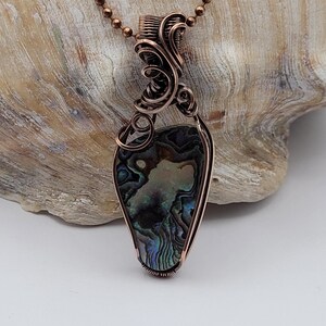 May include: A copper-colored wire-wrapped pendant necklace featuring an abalone shell. The shell displays iridescent blues, greens, and purples. The pendant is suspended from a matching copper-colored chain, creating a unique jewelry piece.