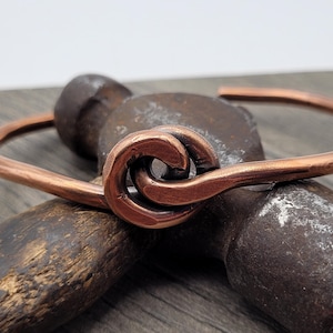 May include: A copper cuff bracelet with a unique knot design. The bracelet is a warm, reddish-brown color and appears to be handmade. The bracelet is resting on a vintage hammer, suggesting a handcrafted or artisan product.