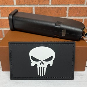 May include: A black patch with a white skull and crossbones design. The skull has a menacing expression with a furrowed brow and a wide, toothy grin. The patch is stitched with a black thread around the edges.