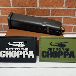May include: Two black and white embroidered patches with the text "Get to the Choppa" and a helicopter silhouette. The patches are on a brown surface. One patch is black with white text and the other is green with black text.