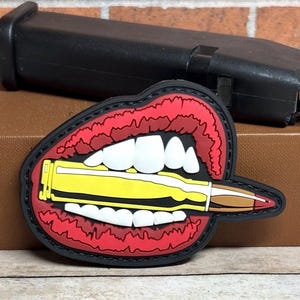 May include: A rubberized morale patch featuring a graphic design of bright red lips with white teeth biting down on a yellow and copper-colored rifle cartridge. The patch has a black border and is shaped to follow the contour of the open mouth design.
