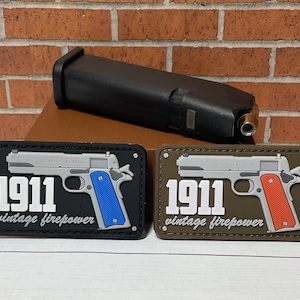 May include: Two black and brown patches with a silver 1911 pistol design. The text "1911 vintage firepower" is printed on each patch. The patches are on a brown wooden surface.