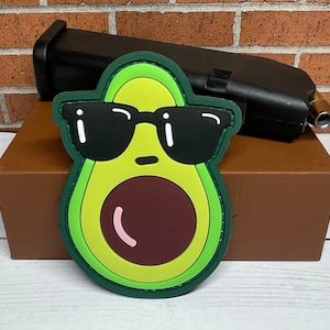 May include: A green avocado with a brown pit and sunglasses. The avocado is wearing a green and black patch with a white outline.