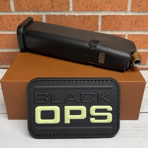May include: A black plastic magazine with a gold-colored bullet tip, sitting on a brown box with a black patch that reads "BLACK OPS" in white letters.
