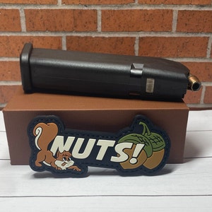 May include: A brown box with a black magazine on top. The box has a black and green patch with a cartoon squirrel and the text "NUTS!" on it.