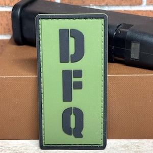 May include: A rectangular olive green patch with the letters "DFQ" in black stencil font. The patch has a black border and is set against a blurred background.